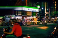 A lively nighttime street scene in front of a FamilyMart convenience store. Neon lights illuminate the area, with several food stalls lined up offering various local dishes. People are seen riding scooters and walking around, contributing to the bustling atmosphere.