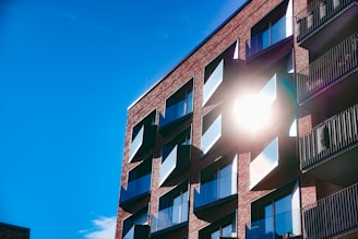 A modern apartment building with a brick facade, featuring large windows and multiple balconies. The sun is shining brightly, reflecting off the windows and casting a stark light on the structure. The sky is clear and vividly blue.