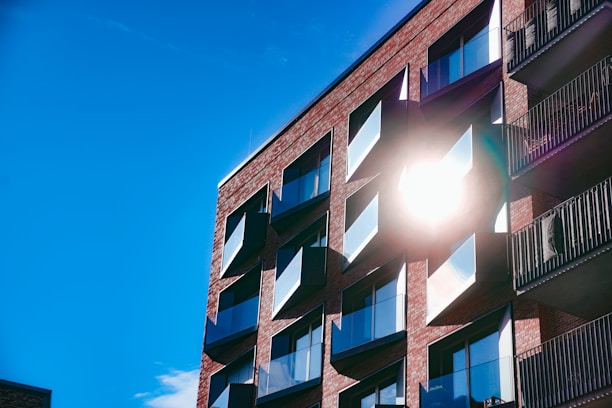 A modern apartment building with a brick facade, featuring large windows and multiple balconies. The sun is shining brightly, reflecting off the windows and casting a stark light on the structure. The sky is clear and vividly blue.