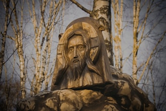 A wooden sculpture depicting a bearded man's face, embedded in a tree-like structure. The expression is solemn and contemplative, surrounded by a background of bare trees with light and shadow interplay, creating an earthy, natural atmosphere.