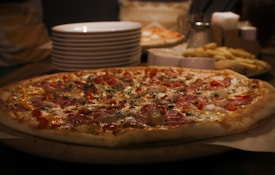 A large pizza topped with slices of ham, chicken, and bell peppers is placed on a wooden table. In the background, several stacks of white plates, a container with utensils, and a serving of fries in a basket are visible. The scene is dimly lit, creating a cozy ambiance.