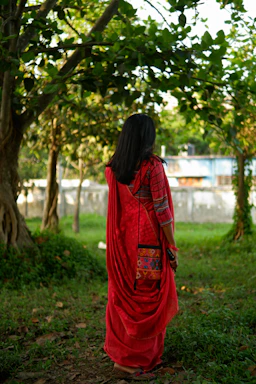 A graceful woman wearing a vibrant red and gold saree, standing in a sunlit garden.