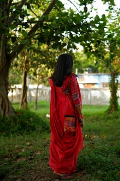 A person wearing a vibrant red saree is standing in a lush, green outdoor setting with tall trees that have dense foliage. The scene suggests a serene and natural environment, possibly a park or garden.