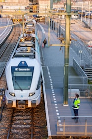 A passenger train is stationed at a platform in a railway station. Two people are visible: one is a railway worker wearing a bright high-visibility jacket and standing on the platform, and the other is a person holding bags, possibly waiting to board. The station has several tracks and platforms with overhead electric lines, and the scene is well-lit with sunlight.