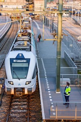 A passenger train is stationed at a platform in a railway station. Two people are visible: one is a railway worker wearing a bright high-visibility jacket and standing on the platform, and the other is a person holding bags, possibly waiting to board. The station has several tracks and platforms with overhead electric lines, and the scene is well-lit with sunlight.