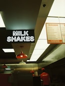 Inside a fast food restaurant with a focus on the milkshake signage. The view includes parts of the kitchen area with two employees in uniforms. Overhead lighting and menu boards are visible.