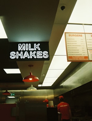 Inside a fast food restaurant with a focus on the milkshake signage. The view includes parts of the kitchen area with two employees in uniforms. Overhead lighting and menu boards are visible.