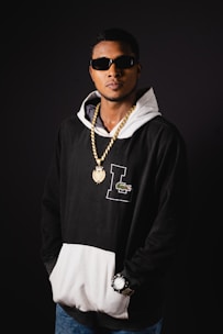 A person wearing sunglasses, a black and white hoodie, and blue jeans. The individual has a gold chain with a pendant and a large watch on their wrist.