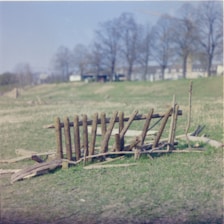A rustic makeshift wooden fence constructed from uneven planks is set against an open field. The background includes a line of bare trees and a few distant buildings under a pale blue sky.