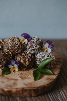A wooden board holding several chocolate truffles coated with chopped nuts and coconut flakes, adorned with purple and white edible flowers and green leaves.
