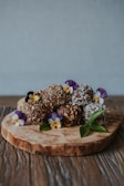 A wooden slab serves as a rustic platter for an assortment of chocolate truffles, generously coated in chopped nuts and coconut flakes. Purple and yellow edible flowers, interspersed between the truffles, add a touch of vibrant color along with a few green leaves for a natural accent. The background is a soft gray, enhancing the earthy tones of the wooden table below.
