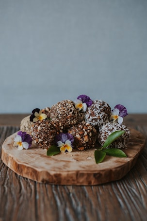 A wooden slab serves as a rustic platter for an assortment of chocolate truffles, generously coated in chopped nuts and coconut flakes. Purple and yellow edible flowers, interspersed between the truffles, add a touch of vibrant color along with a few green leaves for a natural accent. The background is a soft gray, enhancing the earthy tones of the wooden table below.