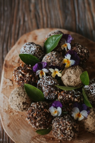 A selection of chocolate truffles is elegantly arranged on a wooden board. The truffles are coated with different toppings such as shredded coconut, chopped nuts, and cocoa powder. Garnishing the arrangement are vibrant green leaves and delicate, colorful flowers which add a fresh and artistic touch to the presentation.