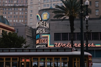 An urban street scene featuring a vintage Walgreens sign with neon lights. The architecture includes a mix of modern and older buildings, framed by palm trees. The foreground shows part of a streetcar, adding to the city's historic ambiance.