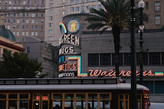 An urban street scene featuring a vintage Walgreens sign with neon lights. The architecture includes a mix of modern and older buildings, framed by palm trees. The foreground shows part of a streetcar, adding to the city's historic ambiance.