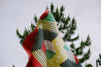 A vibrantly colored, patterned fabric is draped against the backdrop of a cloudy sky and lush green pine tree branches. The fabric features an abstract design with geometric shapes and polka dots in red, yellow, black, white, and green.