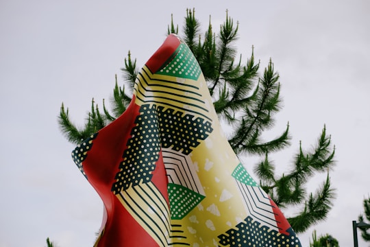 A vibrantly colored, patterned fabric is draped against the backdrop of a cloudy sky and lush green pine tree branches. The fabric features an abstract design with geometric shapes and polka dots in red, yellow, black, white, and green.