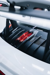 Close-up of a car's rear engine cover with a sleek, modern design, featuring red lights and black vented grilles over a white surface.