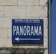 A blue sign on a brick wall with the words 'TOURING-CLUB DE FRANCE' in small letters and 'PANORAMA' in large letters, accompanied by an arrow pointing left.