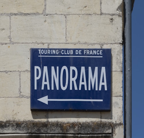 A blue sign on a brick wall with the words 'TOURING-CLUB DE FRANCE' in small letters and 'PANORAMA' in large letters, accompanied by an arrow pointing left.