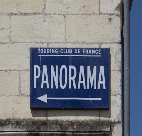 A blue sign on a brick wall with the words 'TOURING-CLUB DE FRANCE' in small letters and 'PANORAMA' in large letters, accompanied by an arrow pointing left.