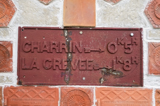 A vintage, weathered metal plaque mounted on a stone wall. The plaque features raised text in French, indicating directional information, along with numerical details. Surrounding the plaque are decorative terracotta tiles, each embossed with intricate patterns and text.