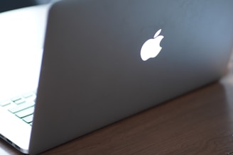 A laptop with a glowing logo on the back of the screen, placed on a wooden surface. The focus is on the illuminated logo, and the keyboard is partially visible.