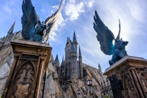 A grand, magical castle with towering spires set against a clear blue sky. Two large stone statues of winged creatures stand atop ornate pedestals at the entrance, framed by rocky terrain and decorative lighting.