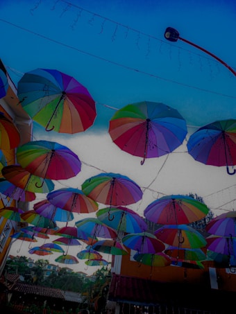 Multiple colorful umbrellas are suspended in the air, creating a vibrant canopy in an outdoor setting. The umbrellas are arranged closely together, hanging by strings, against a picturesque backdrop of buildings and a clear blue sky. Fairy lights are also strung alongside the umbrellas, adding a whimsical touch.