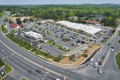 Aerial view of a commercial shopping area featuring a large parking lot filled with numerous cars. Several retail stores and a restaurant are visible, surrounded by greenery and bordered by two main roads with multiple vehicles on them. A few trees and shrubs are interspersed within the parking area.