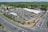Aerial view of a commercial shopping area featuring a large parking lot filled with numerous cars. Several retail stores and a restaurant are visible, surrounded by greenery and bordered by two main roads with multiple vehicles on them. A few trees and shrubs are interspersed within the parking area.