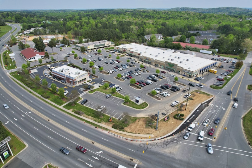 Aerial view of a commercial shopping area featuring a large parking lot filled with numerous cars. Several retail stores and a restaurant are visible, surrounded by greenery and bordered by two main roads with multiple vehicles on them. A few trees and shrubs are interspersed within the parking area.