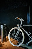 A sleek, shiny bicycle with a chrome finish is positioned against a dark background. The handlebars and a part of the frame are highlighted, with white tires standing out against a textured brown surface beneath.