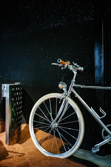 A sleek, shiny bicycle with a chrome finish is positioned against a dark background. The handlebars and a part of the frame are highlighted, with white tires standing out against a textured brown surface beneath.