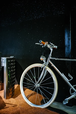 A sleek, shiny bicycle with a chrome finish is positioned against a dark background. The handlebars and a part of the frame are highlighted, with white tires standing out against a textured brown surface beneath.