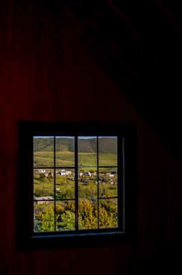 A picturesque rural landscape is visible through a window, featuring lush green fields, scattered houses, and rolling hills in the background. The view is framed by a dark, rustic interior, creating a contrast between the dimly lit room and the vibrant scenery outside.