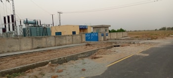 A power station with electrical transformers and equipment enclosed within a boundary wall. Signs with text are visible on the blue gate and walls. The facility is situated next to a road with open fields in the background under a clear sky.