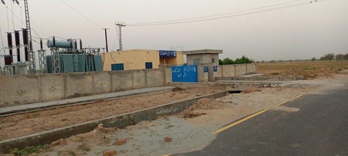 A power station with electrical transformers and equipment enclosed within a boundary wall. Signs with text are visible on the blue gate and walls. The facility is situated next to a road with open fields in the background under a clear sky.