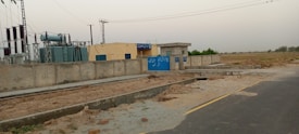 A power station with electrical transformers and equipment enclosed within a boundary wall. Signs with text are visible on the blue gate and walls. The facility is situated next to a road with open fields in the background under a clear sky.