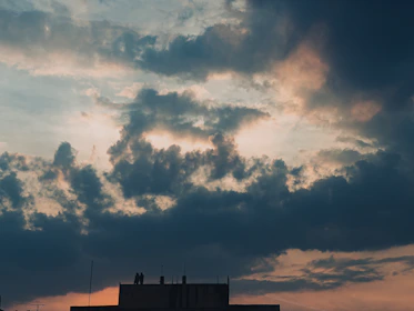An atmospheric shot of a dimly lit urban rooftop at night, with silhouettes of two figures cloaked in shadows under a moody sky.