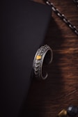 A silver bracelet with intricate, scale-like patterns and a small golden accent resting on the edge of a black, textured surface. The background includes a wooden surface and part of a chain in the upper right corner.