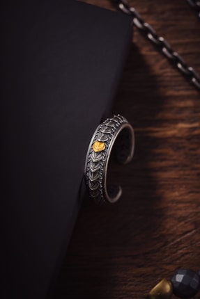 A silver bracelet with intricate, scale-like patterns and a small golden accent resting on the edge of a black, textured surface. The background includes a wooden surface and part of a chain in the upper right corner.
