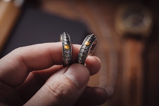 A close-up of a hand holding two intricately designed rings with yellow gemstones. The rings have a detailed pattern and are silver in color. The background appears blurred, suggesting focus on the jewelry pieces.