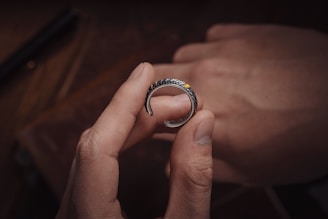 A hand delicately holds a silver ring with intricate carvings and a small amber gemstone. Another hand is visible in the background, slightly out of focus. The background features a warm wooden texture, adding an earthy feel.