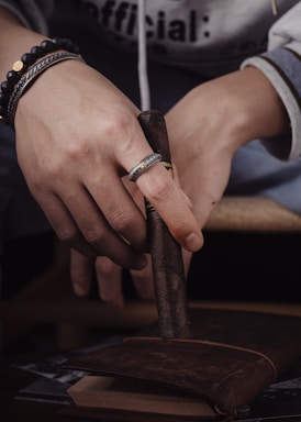 A person's hands holding a brown cigar above a leather-bound book. The person is wearing a ring with an intricate design and several bracelets, including a black beaded one.