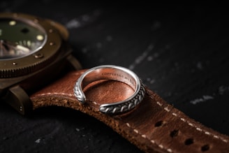 A close-up view of a silver ring with intricate patterns placed on a brown leather watch strap. The ring has an inscription on the inside, and the watch is partially visible with a metallic casing. The surface underneath is dark and textured.