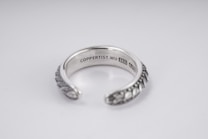 A silver open-ended ring with intricate patterns on the outer surface. The inside is engraved with the text 'COPPERTIST.WU S925 ANT' indicating sterling silver. The design is elegant and detailed, complemented by a smooth inner finish.