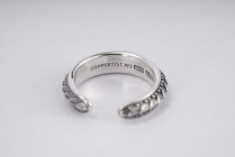 A silver open-ended ring with intricate patterns on the outer surface. The inside is engraved with the text 'COPPERTIST.WU S925 ANT' indicating sterling silver. The design is elegant and detailed, complemented by a smooth inner finish.