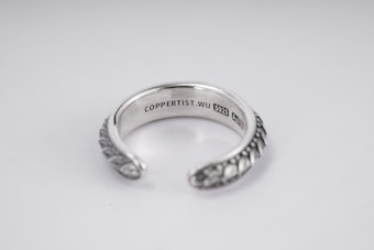 A silver open-ended ring with intricate patterns on the outer surface. The inside is engraved with the text 'COPPERTIST.WU S925 ANT' indicating sterling silver. The design is elegant and detailed, complemented by a smooth inner finish.