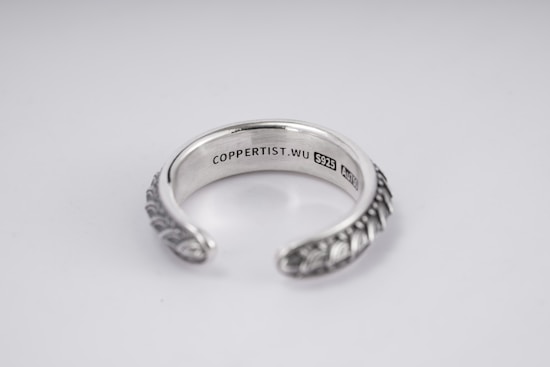 A silver open-ended ring with intricate patterns on the outer surface. The inside is engraved with the text 'COPPERTIST.WU S925 ANT' indicating sterling silver. The design is elegant and detailed, complemented by a smooth inner finish.